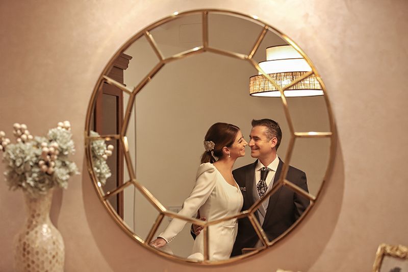 Los novios se miran en un espejo redondo con marco dorado. Se ven la pared rosa, el vestido blanco y el ramo.