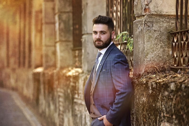 Hombre con traje a cuadros azul apoyado contra un viejo muro de piedra, mirando a la cámara.