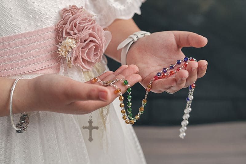 Niño con vestido blanco sosteniendo rosario, detalle de manos con pulseras.