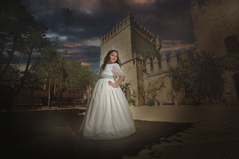 Chica con vestido blanco posa frente al castillo, cielo oscuro.