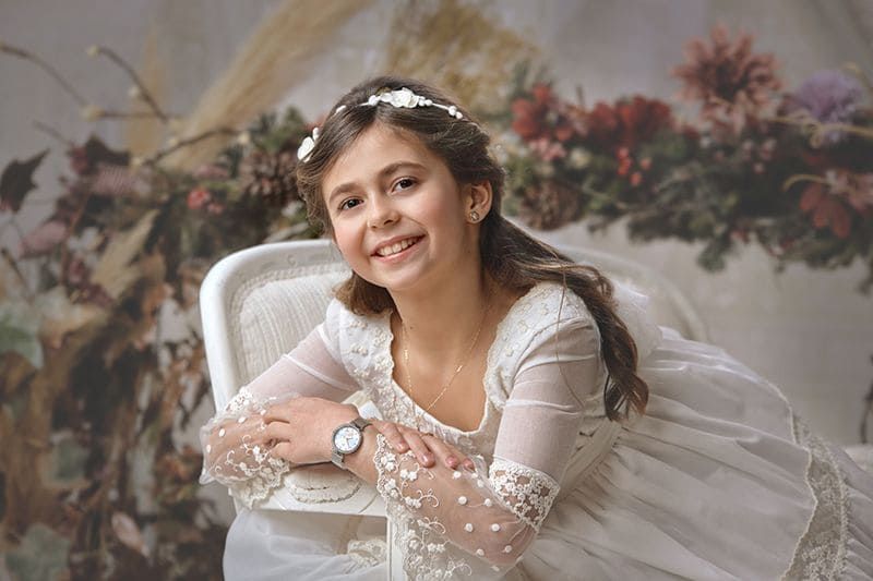 Chica joven con vestido blanco, sonriendo y con una diadema de flores, descansando en una silla blanca. Fondo floral.