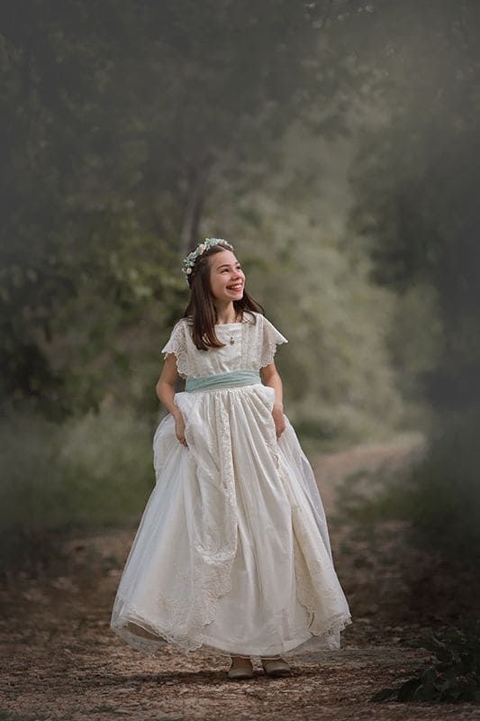 Una joven vestida de blanco y con una faja azul se ríe en un sendero en un bosque, luciendo una corona de flores.