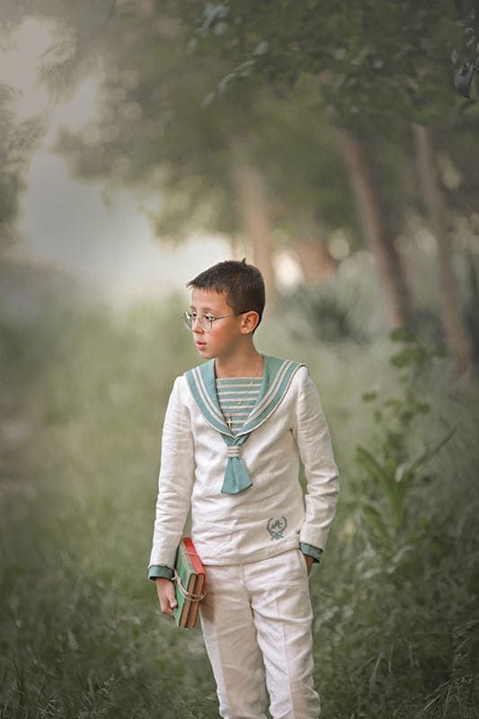 Niño con traje de marinero, gafas, sosteniendo un libro, de pie en un entorno al aire libre de tonos verdes.