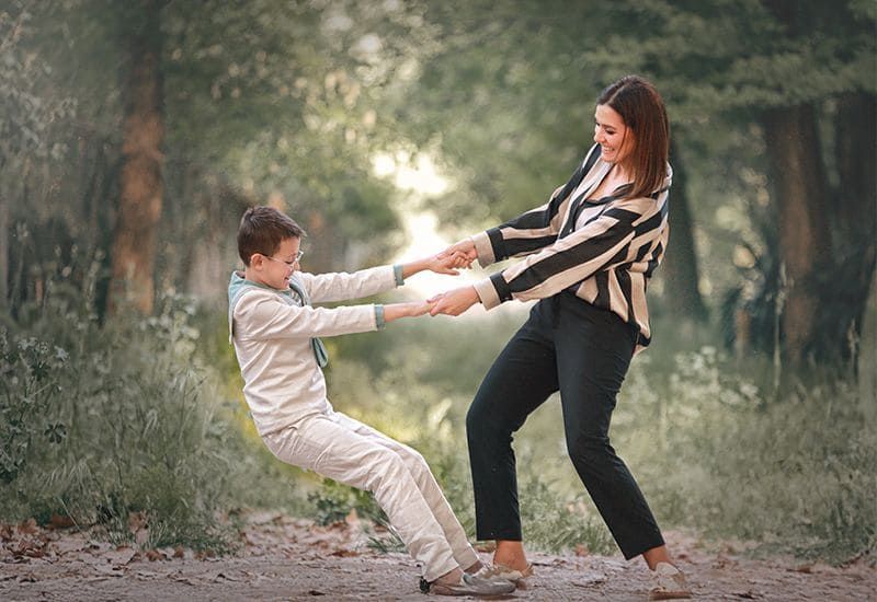 Mujer y niño se toman de la mano, juguetonamente, se jalan al aire libre. Él se recuesta, ella se queda de pie, ambos sonriendo.