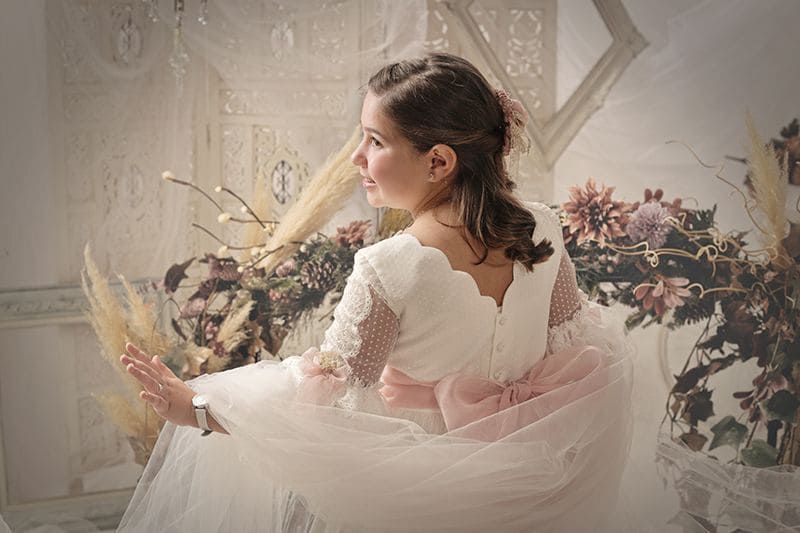 Niña con vestido blanco y lazo rosa, de pie frente a un arreglo floral.