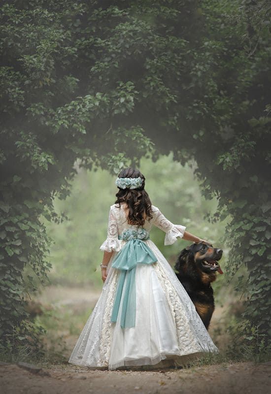 Mujer vestida de blanco con perro, entre vegetación arqueada.