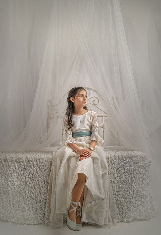 Una chica con vestido blanco y faja azul está sentada en una cama, pensativa. Detrás hay una tela blanca.