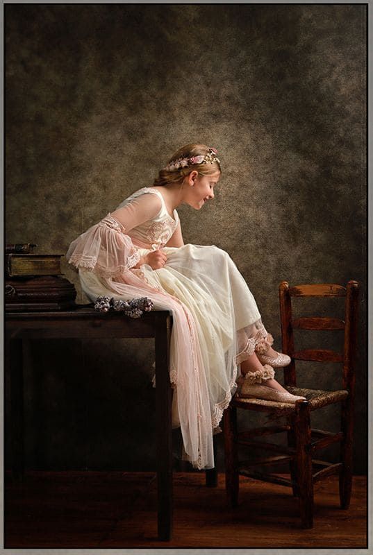 Una joven vestida de blanco se sienta en un escritorio, mirando sus zapatos en una silla de madera, sonriendo.