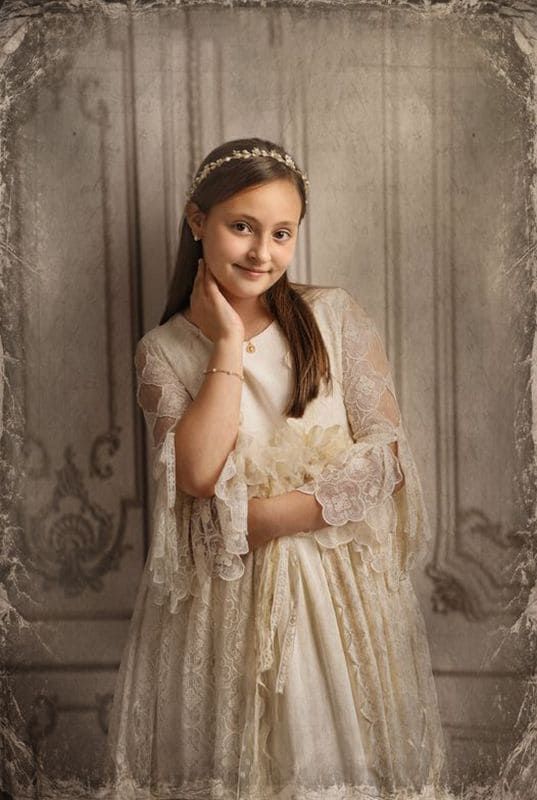 Niña con vestido de encaje blanco y diadema, sonriendo, con la mano en el cuello, frente a una pared adornada.