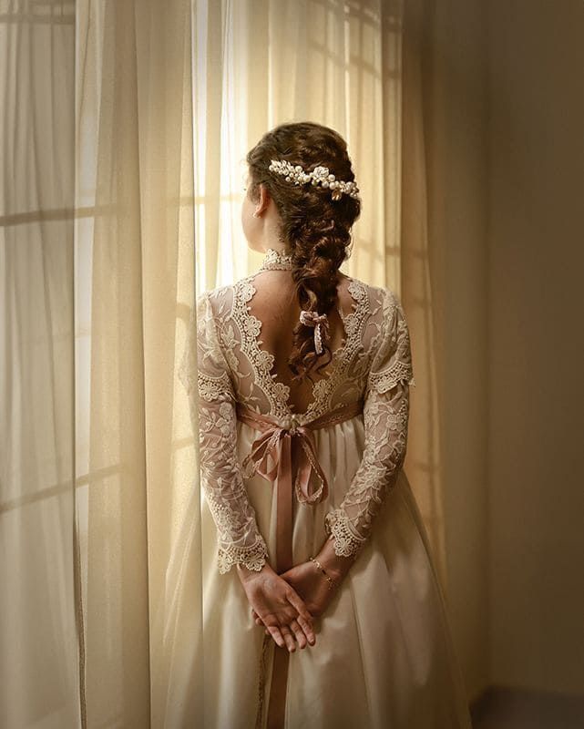 Mujer con vestido de novia mira por la ventana, de espaldas a la cámara. Mangas de encaje, trenza y detalle de cinta.