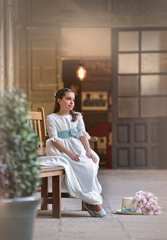 Una chica con vestido blanco y faja azul está sentada en un banco. Un sombrero con flores está en el suelo.