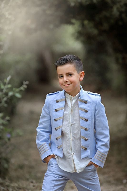 Niño con chaqueta y pantalones estilo militar azul claro, con las manos en los bolsillos, sonriendo al aire libre.