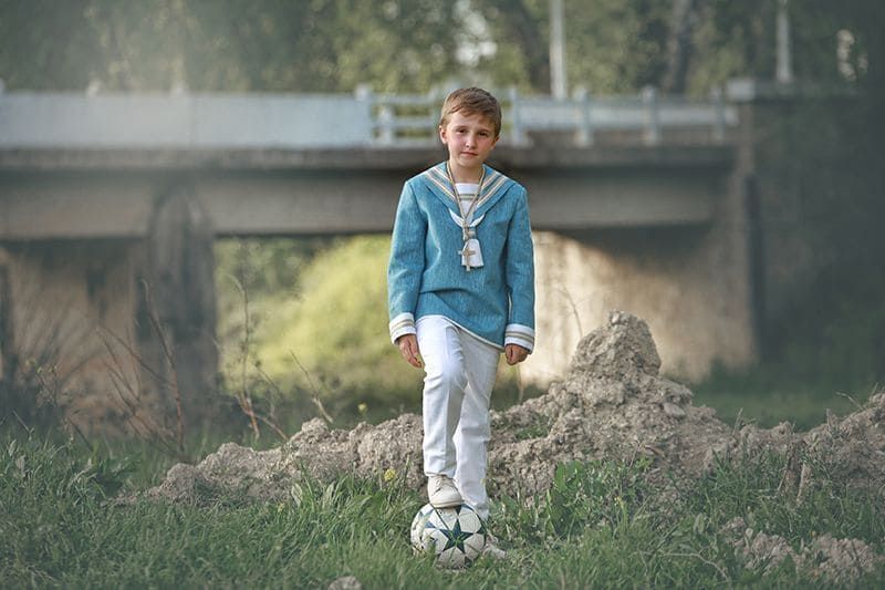 Un niño con camisa de marinero azul y pantalones blancos se encuentra sobre un balón de fútbol frente a un puente.