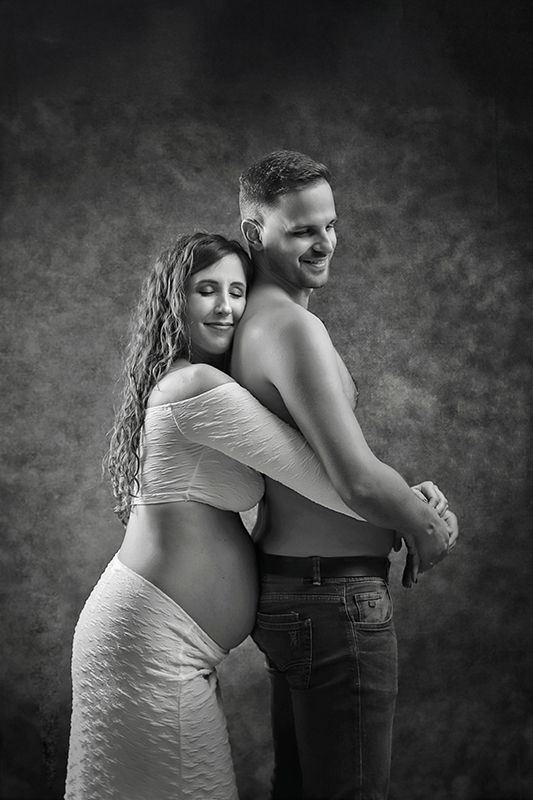 Una mujer embarazada abraza a un hombre sin camisa, ambos sonriendo, contra un fondo oscuro.