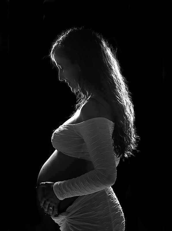 Mujer embarazada en silueta, manos tocando suavemente su vientre, vestido blanco suave, fondo negro.