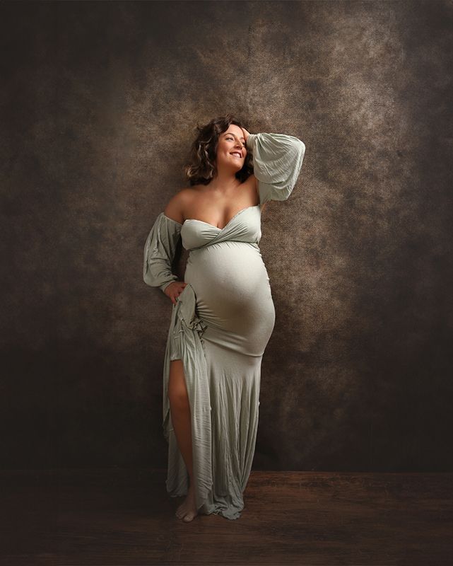 Mujer embarazada con un vestido verde salvia, sonríe, de pie, con la mano en la cabeza, contra un fondo marrón.