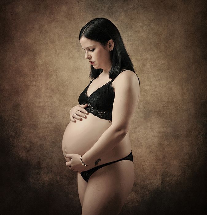 Mujer embarazada en lencería negra, sosteniendo su vientre, mirando hacia abajo, sobre un fondo marrón.