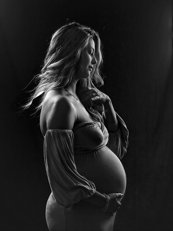 Mujer embarazada de perfil, con el vientre agarrado y vestido con hombros descubiertos. Retrato de estudio en blanco y negro.