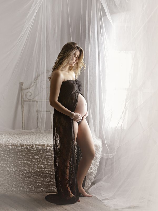 Mujer embarazada con vestido de encaje negro, de pie junto a la ventana, con la mano en el vientre. Luz suave y brillante.