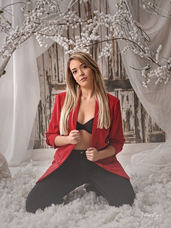 Una mujer con blazer rojo y sujetador negro se arrodilla en un entorno nevado, con un fondo blanco y de madera.