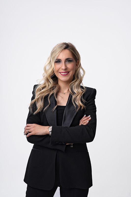 Mujer de cabello rubio, vestida con un blazer negro, con los brazos cruzados, sonríe a la cámara sobre un fondo blanco.