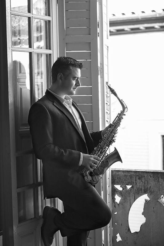 Hombre de traje con un saxofón, apoyado en el marco de una ventana. Blanco y negro.
