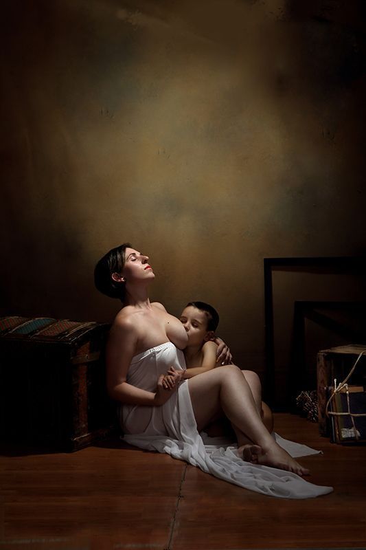 Mujer con bata blanca y un niño, sentada en el suelo, mirando hacia arriba. Escenario oscuro.