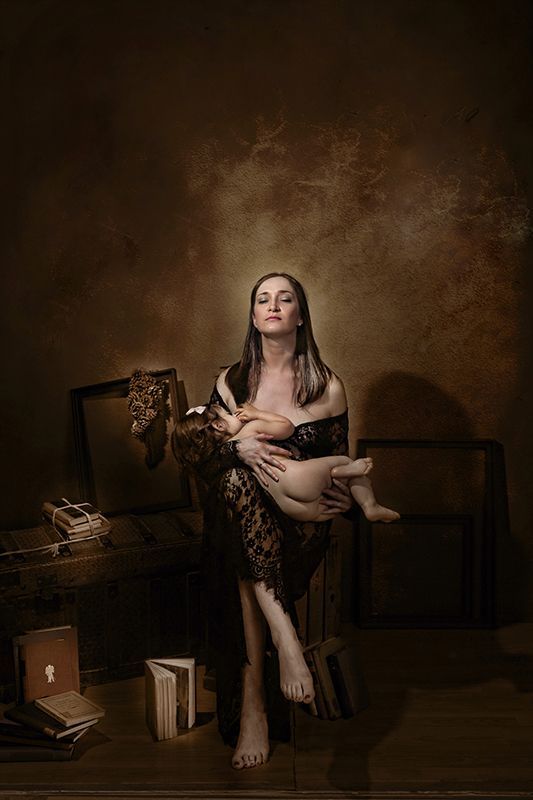 Una mujer amamantando a un bebé, iluminada dramáticamente. Está en una habitación oscura, rodeada de libros y equipaje.