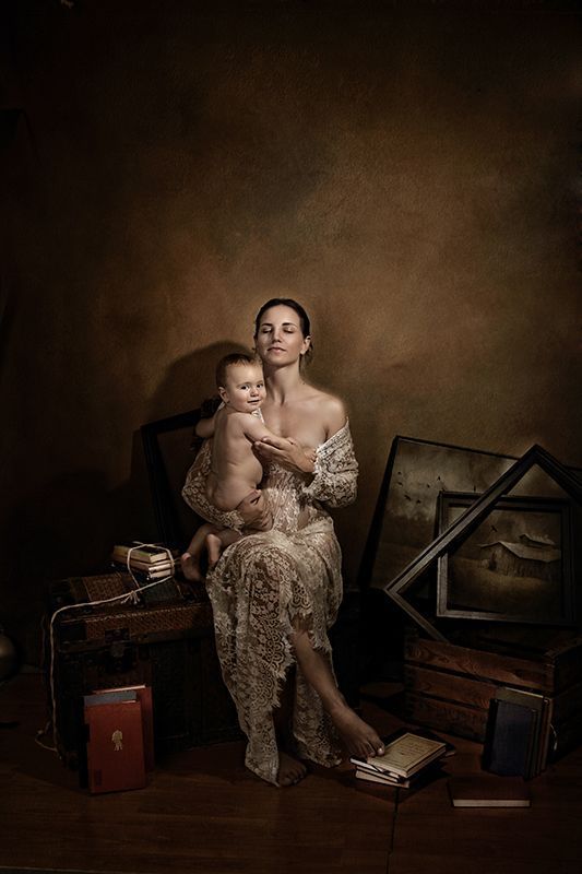 Una mujer con un vestido de encaje acuna a un bebé; sentada en un baúl de madera, contra un fondo marrón.