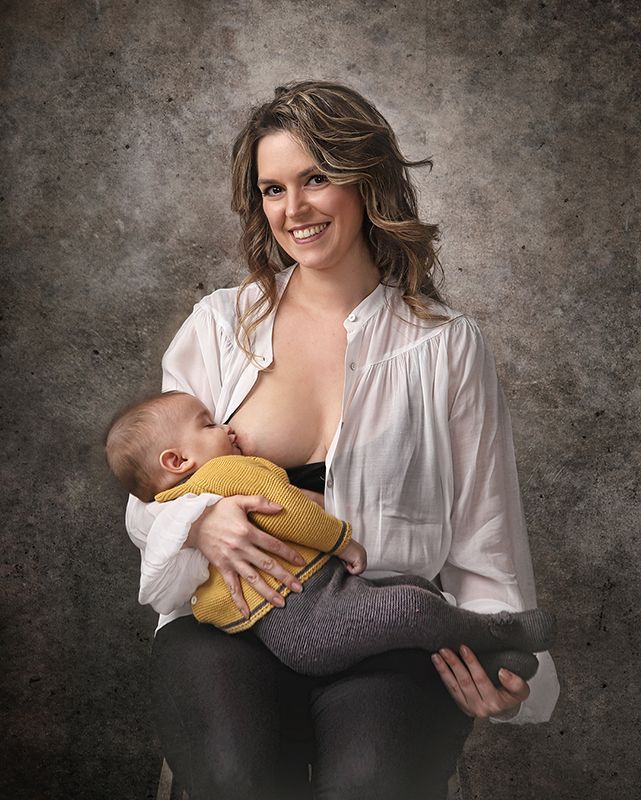 Una mujer sonríe amamantando a su bebé. Sobre un fondo gris texturizado, viste camisa blanca y pantalón negro.