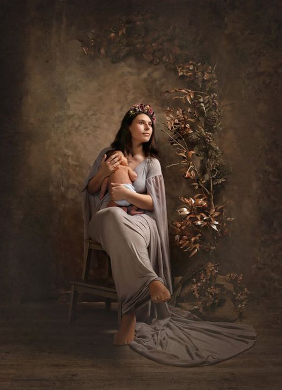 Mujer con un vestido gris fluido sostiene a un bebé con una corona floral, sentada en un taburete rústico. Fondo beige y dorado.