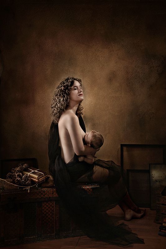 Mujer amamantando a un bebé, envuelta en tela negra, fondo marrón, iluminada desde arriba, mirando hacia arriba.