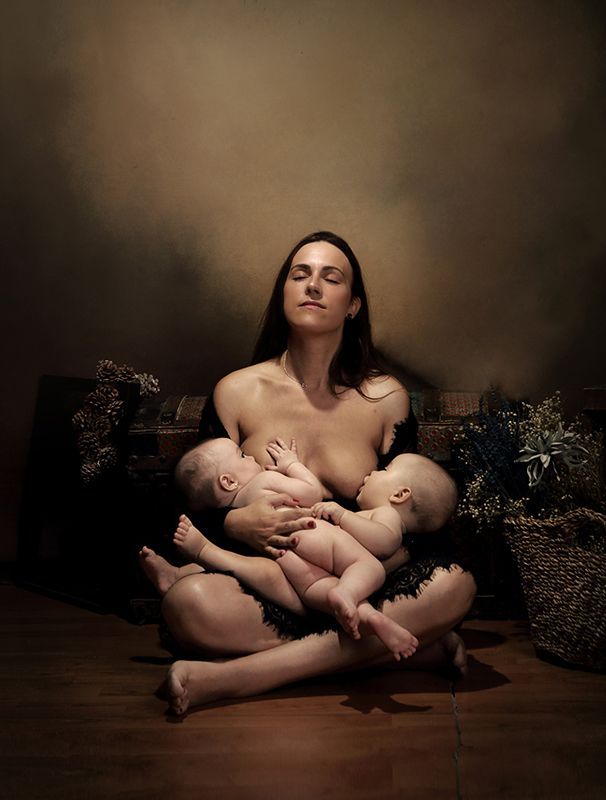 Mujer sentada amamantando a sus gemelos, con los ojos cerrados, contra un fondo marrón poco iluminado.