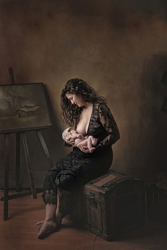 Mujer con vestido de encaje negro amamantando a su bebé, sentada en un baúl cerca de un cuadro.