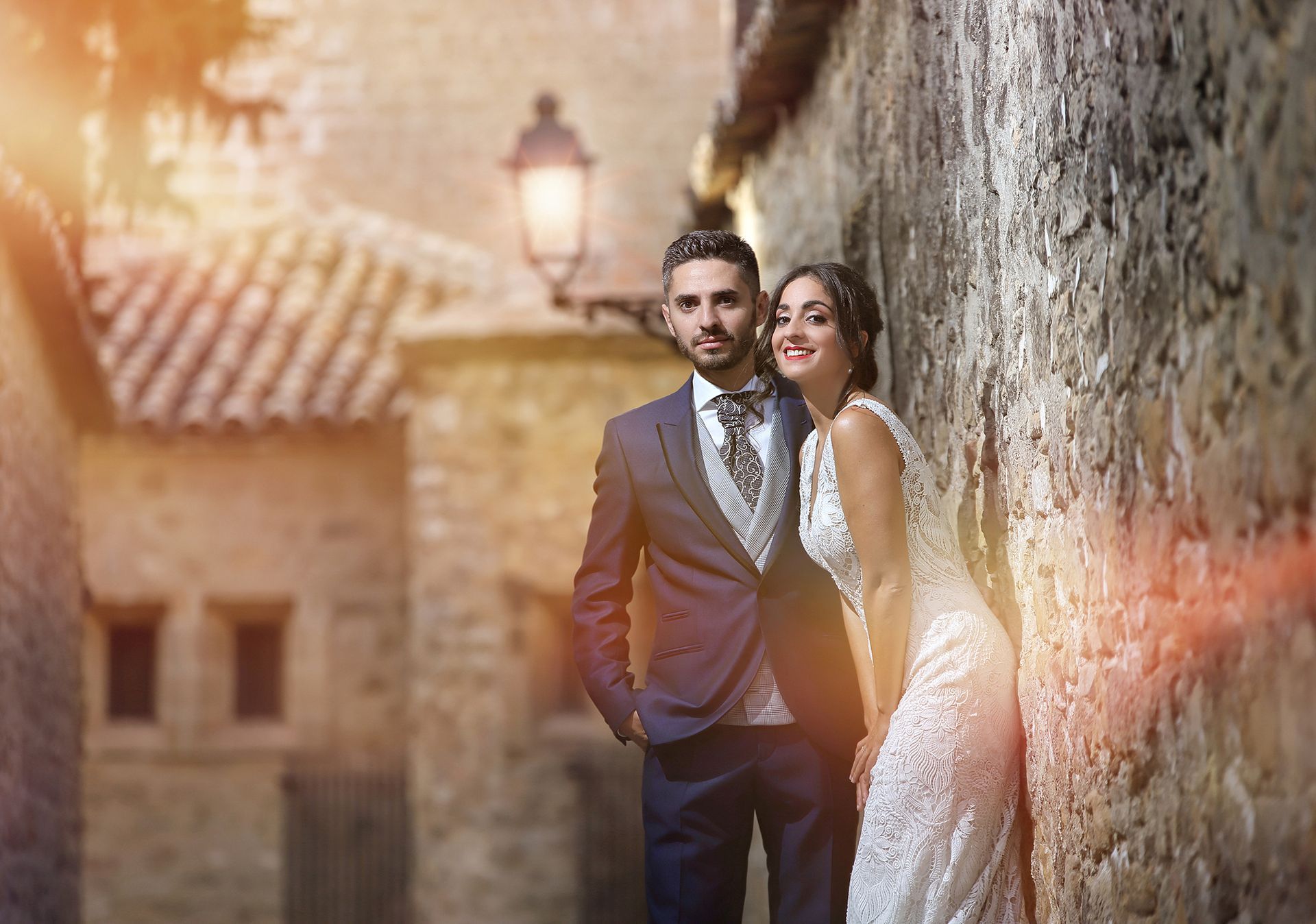 Los novios vestidos de novia posan frente a un muro de piedra en un pueblo antiguo; efecto de destello solar.