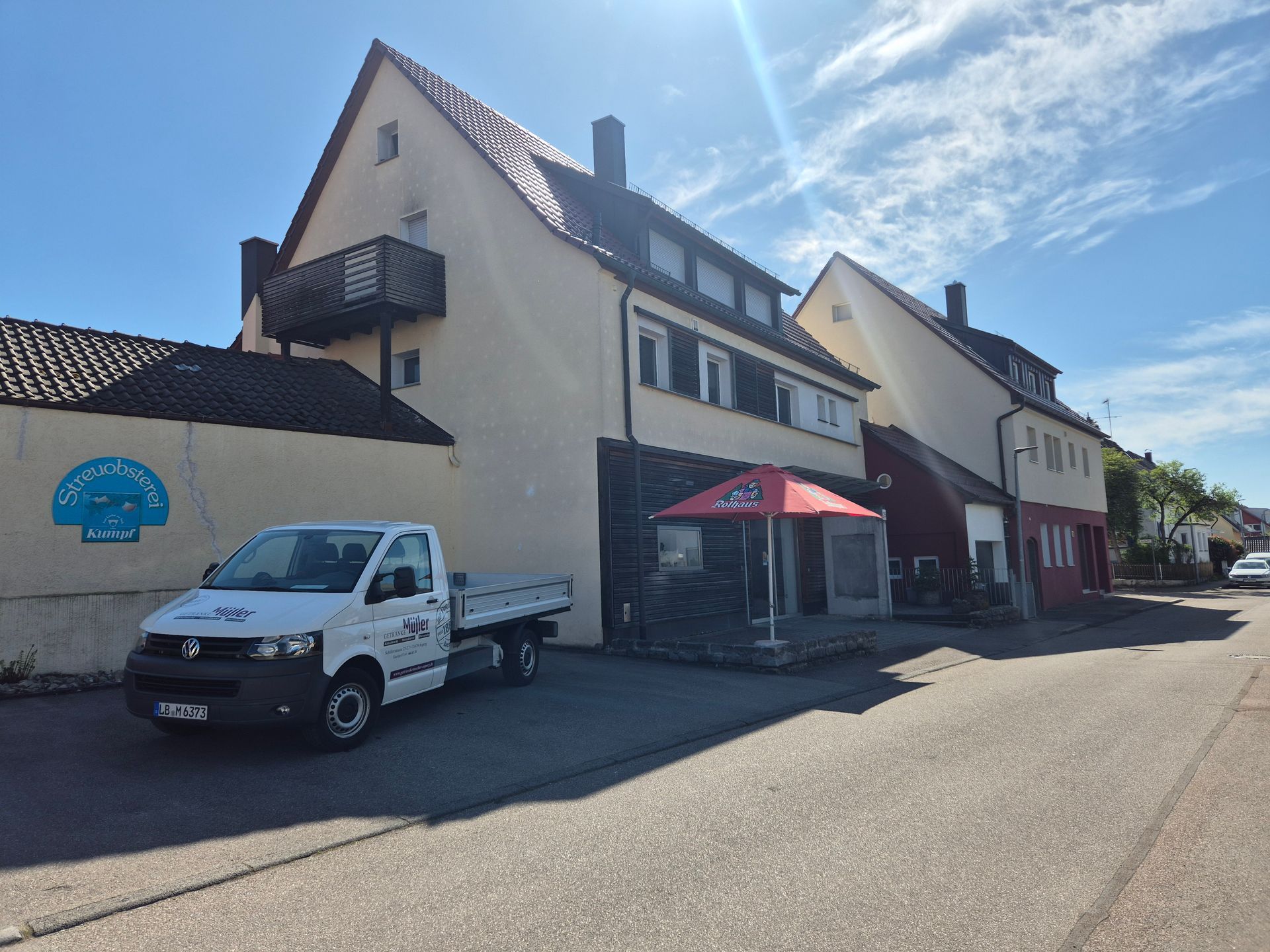 Straße mit weißen Gebäuden, einem geparkten weißen Lieferwagen und Sitzgelegenheiten im Freien unter einem roten Sonnenschirm
