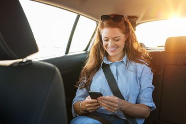 Une femme souriante, assise dans un taxi, qui consulte son téléphone.