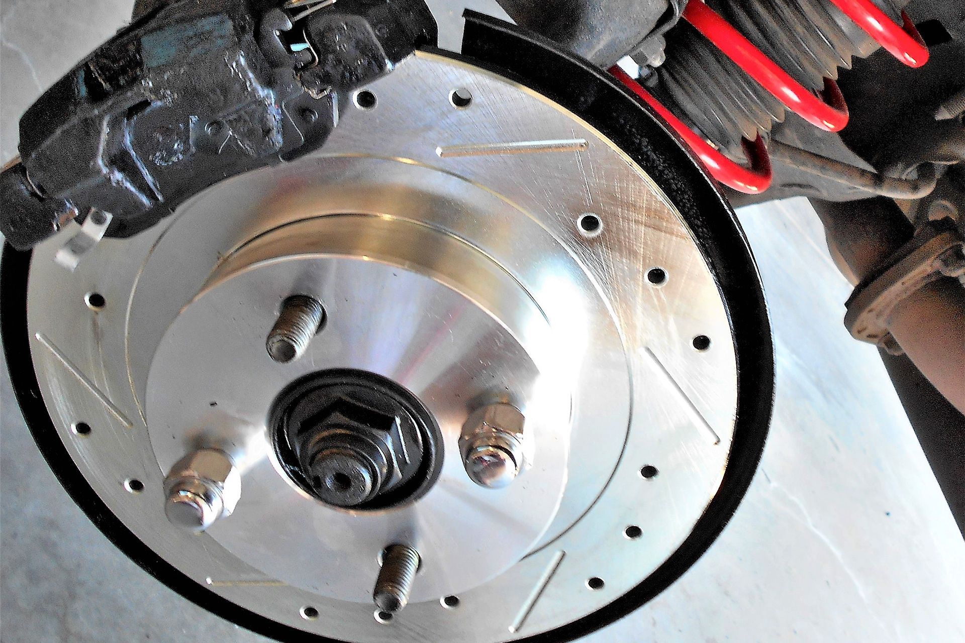 Imagem em close-up de um disco de freio prateado, ranhurado e perfurado, com parafusos pretos, montado no chassi de um carro.