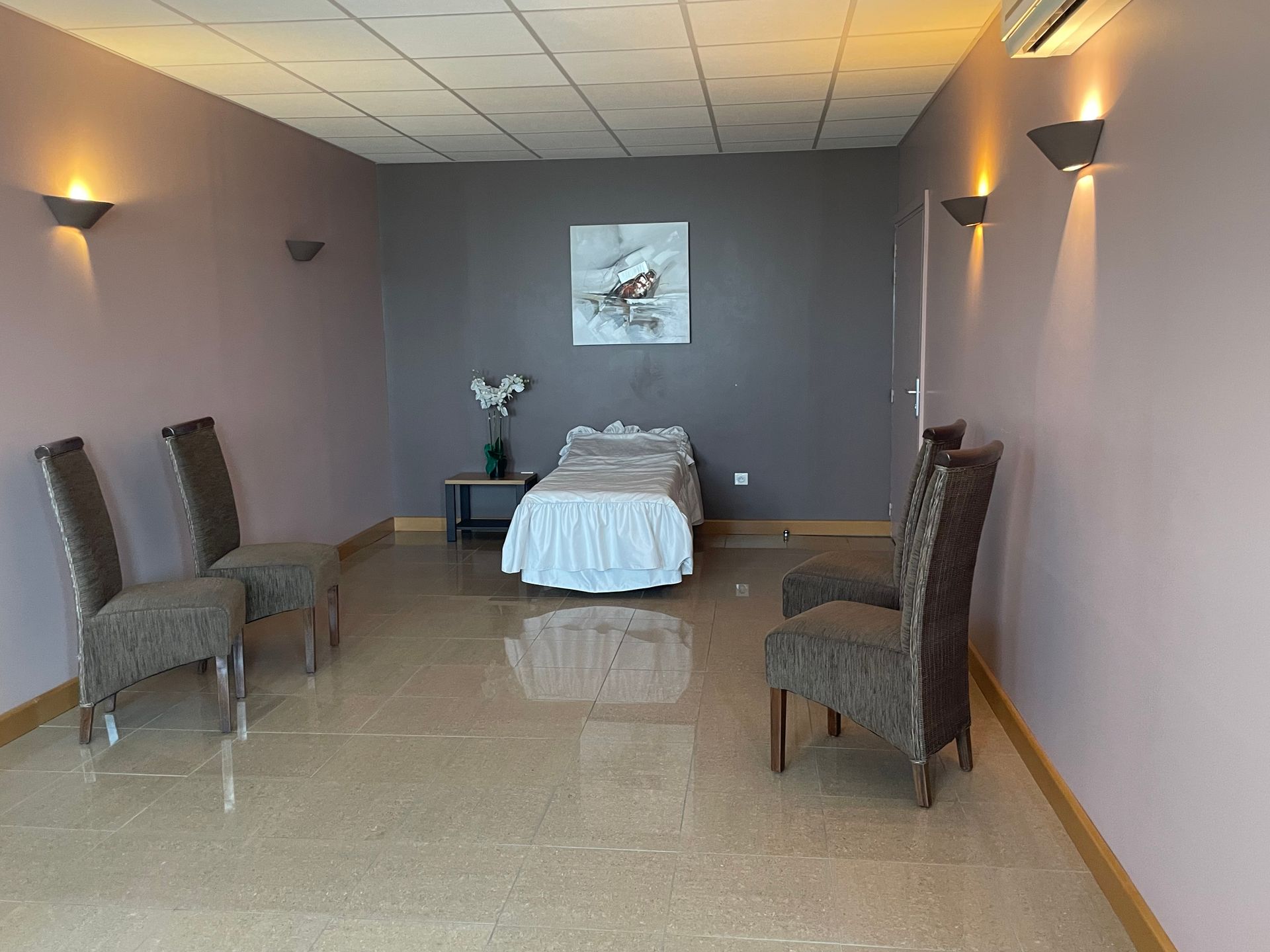 Salle d'attente avec chaises, lit et œuvres d'art. Murs rose tendre, carrelage beige et éclairage chaleureux.