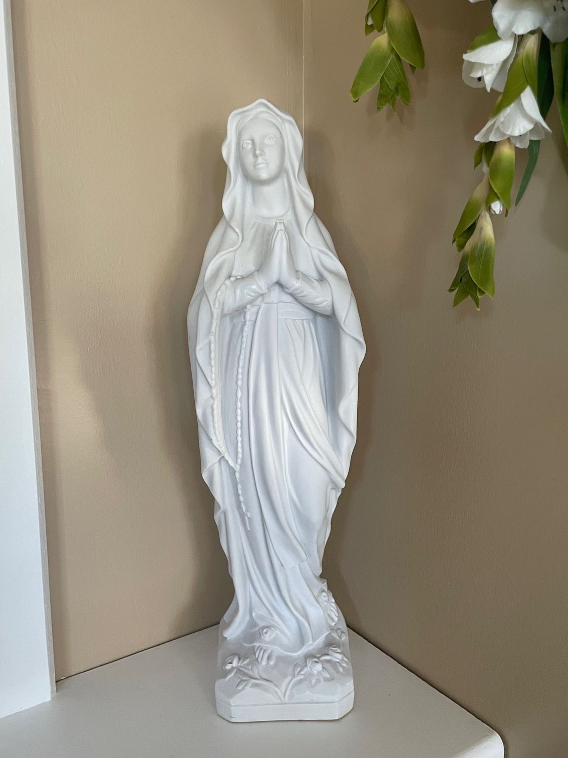 Statue blanche de la Vierge Marie, les mains jointes en prière, debout sur un rebord.