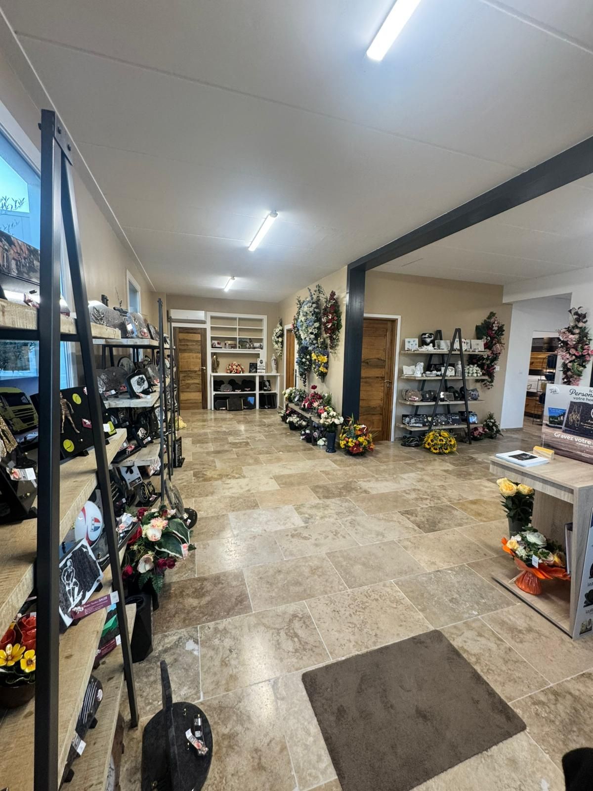 Intérieur d'un fleuriste avec compositions florales, présentoirs et étagères. Carrelage beige, murs neutres et éclairage lumineux.