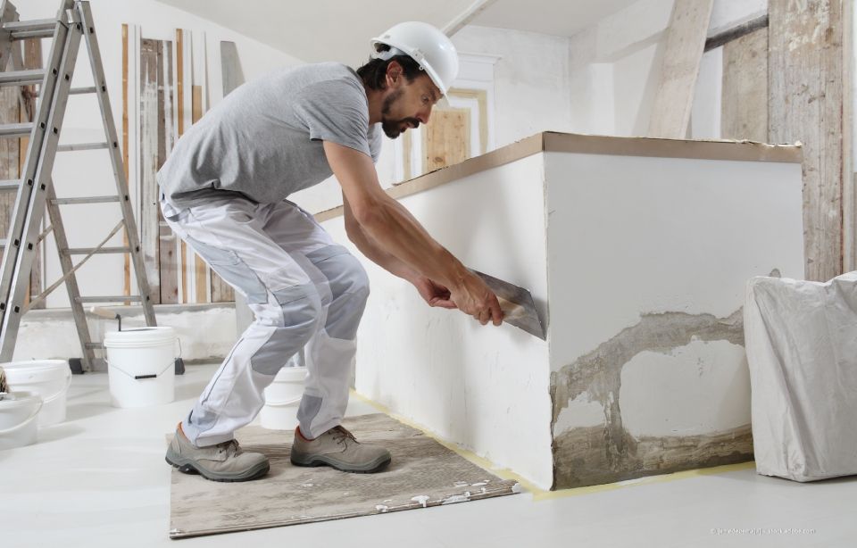 Person trägt mit Handschuhen und einer Kelle Putz auf eine Ziegelwand auf.