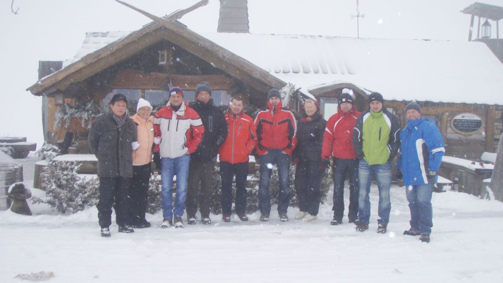 Eine Gruppe von Menschen in Winterkleidung steht vor einem schneebedeckten Holzgebäude. Es schneit.