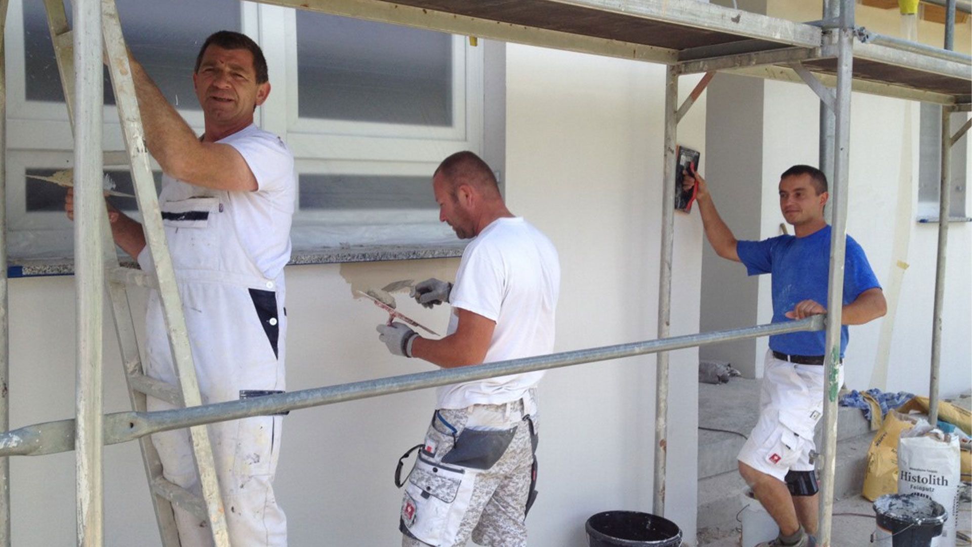 Drei Bauarbeiter verputzen mithilfe eines Gerüsts eine Außenwand.