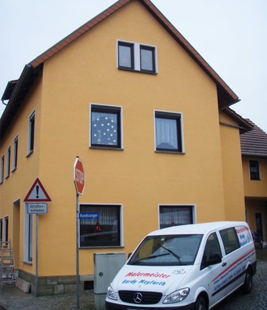 Gelbes Haus mit schwarz gerahmten Fenstern; davor parkt ein weißer Lieferwagen. Straßenschild und Warnsymbol sind ebenfalls zu sehen.