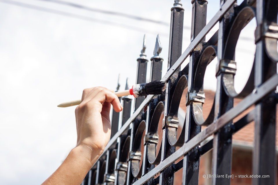 Person, die im Freien einen schwarzen Metallzaun mit einem Pinsel bemalt.