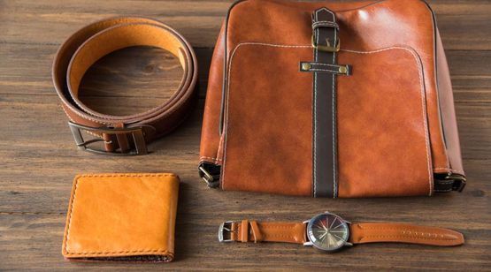 Un bolso de cuero marrón, un cinturón, una billetera y un reloj están sobre una mesa de madera.