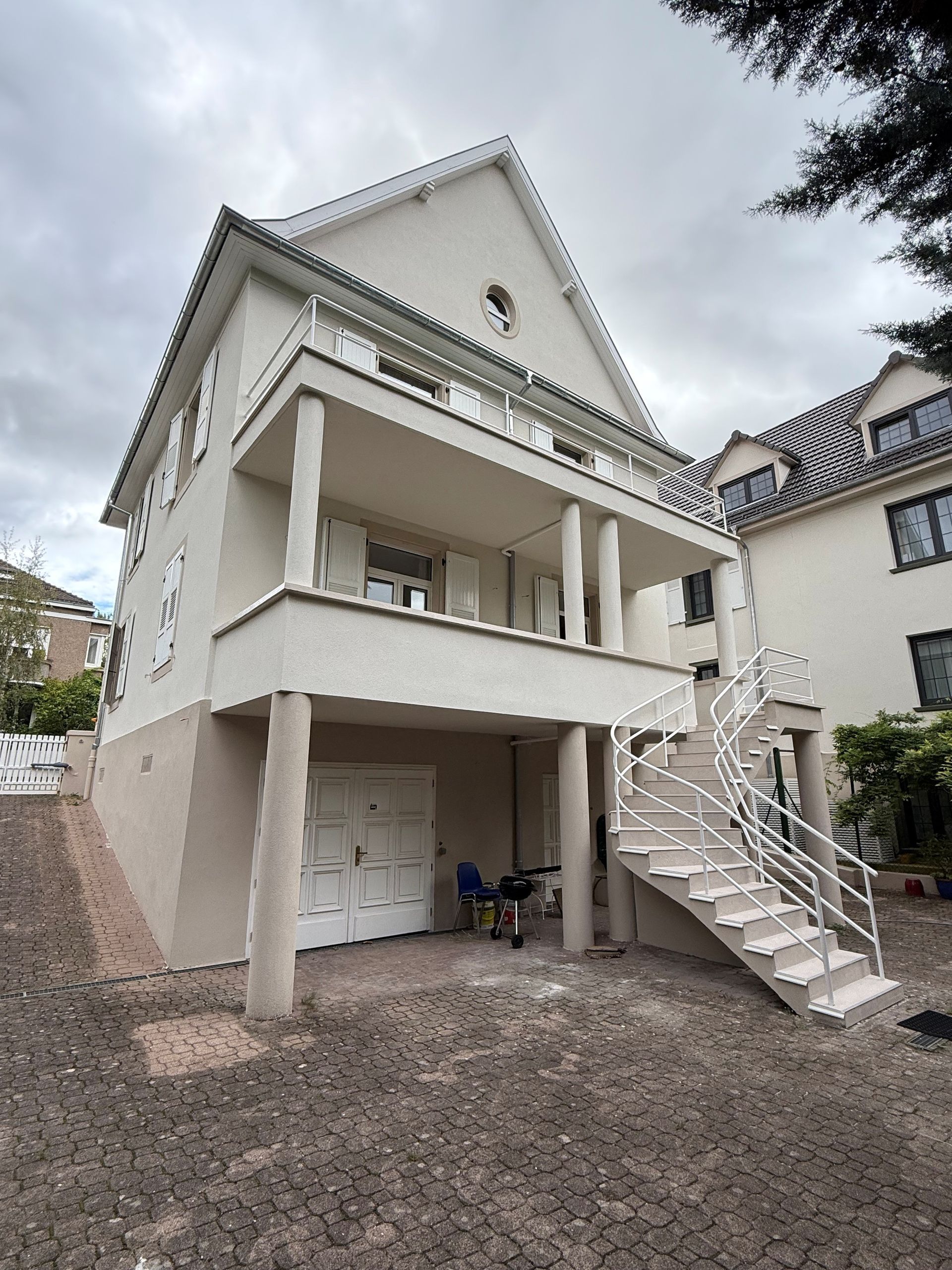 Maison blanche avec toit et balcon sombres, mur en pierre grise, lampadaire.