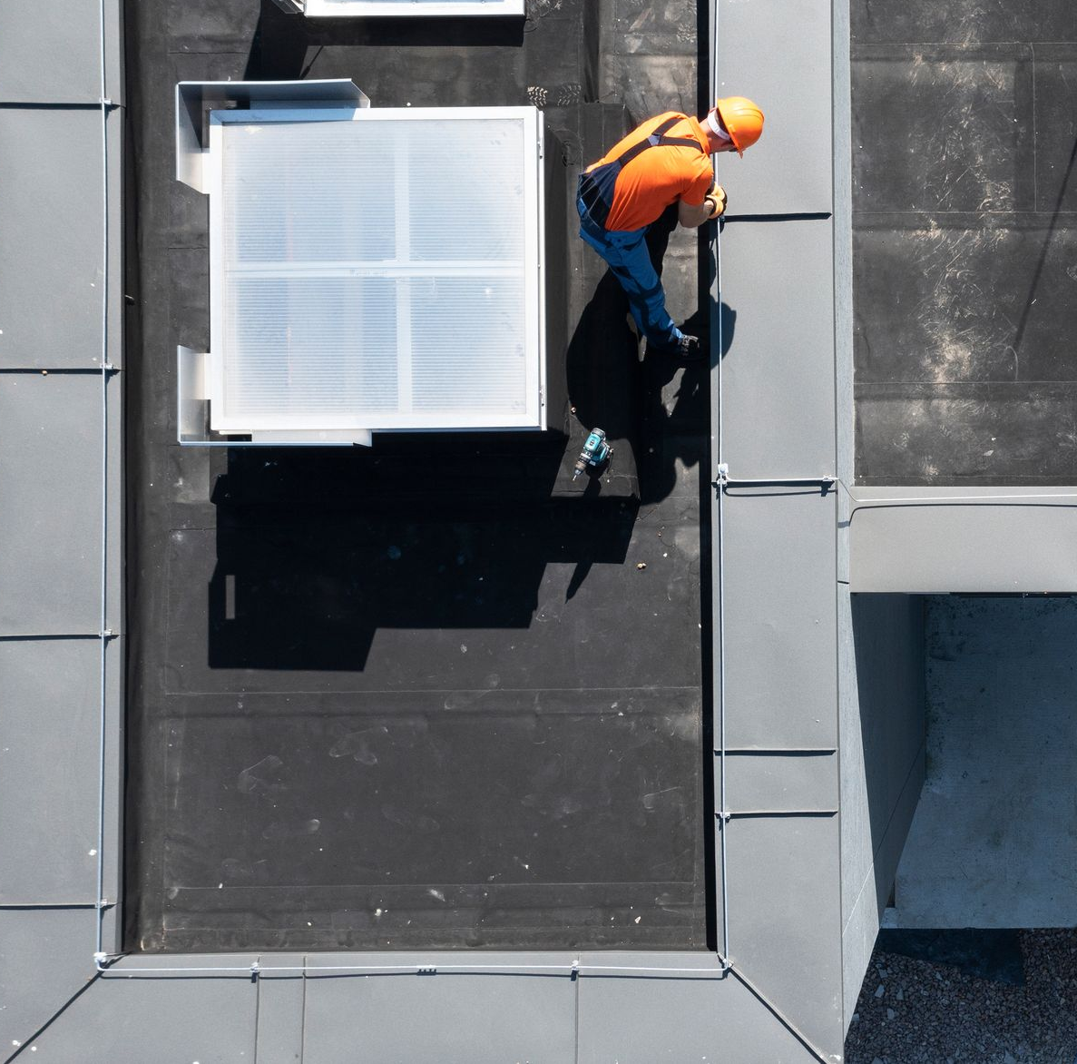 Ouvrier travaillant sur un toit, vêtu d'un gilet orange et d'un casque de chantier, près du bord d'un bâtiment.