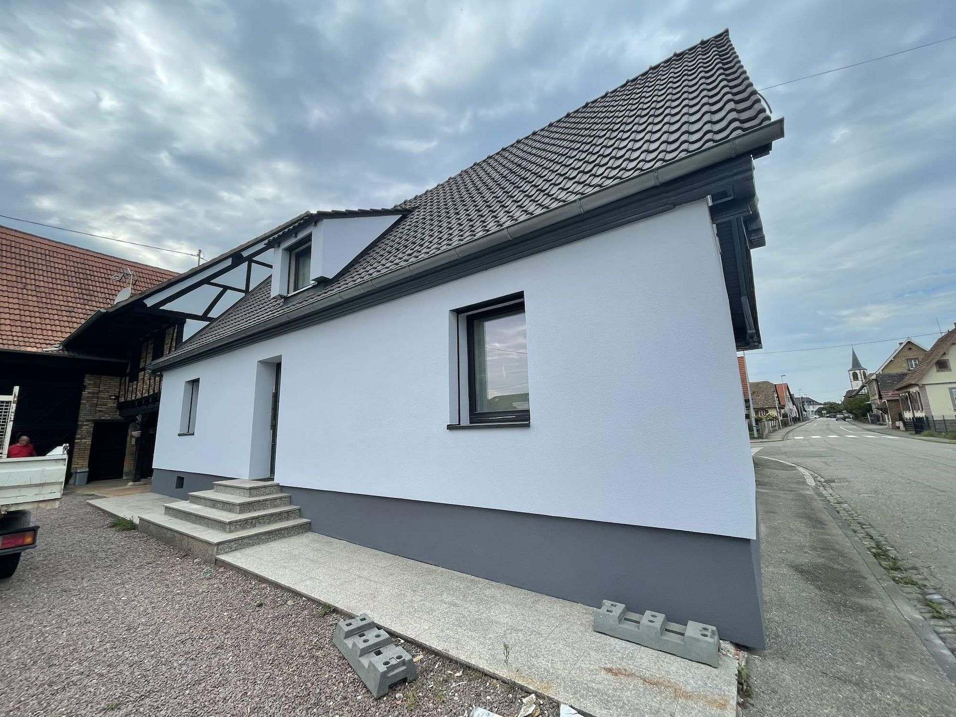 Bâtiment gris et blanc avec un toit et des boiseries gris foncé, sous un ciel nuageux.